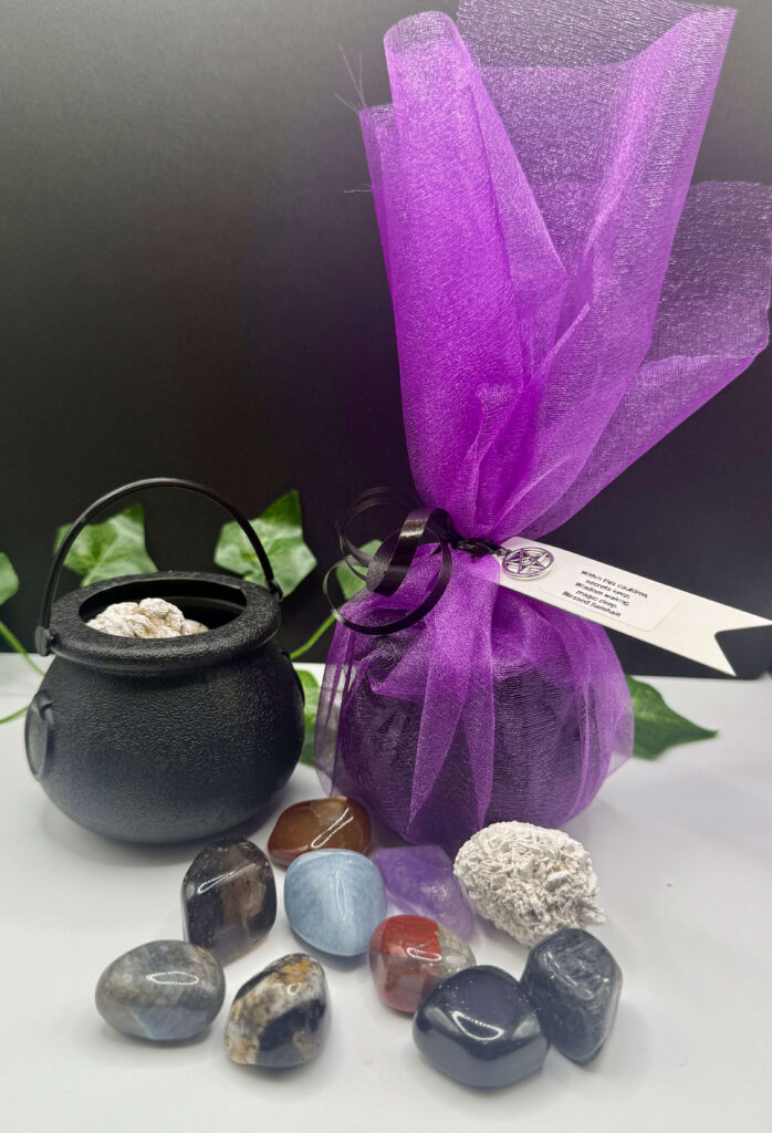 Vibrant purple gift wrap with black ribbon and charm, black cauldron filled with crystals, and assorted polished healing stones with green foliage, creating a mystical and spiritual aesthetic.