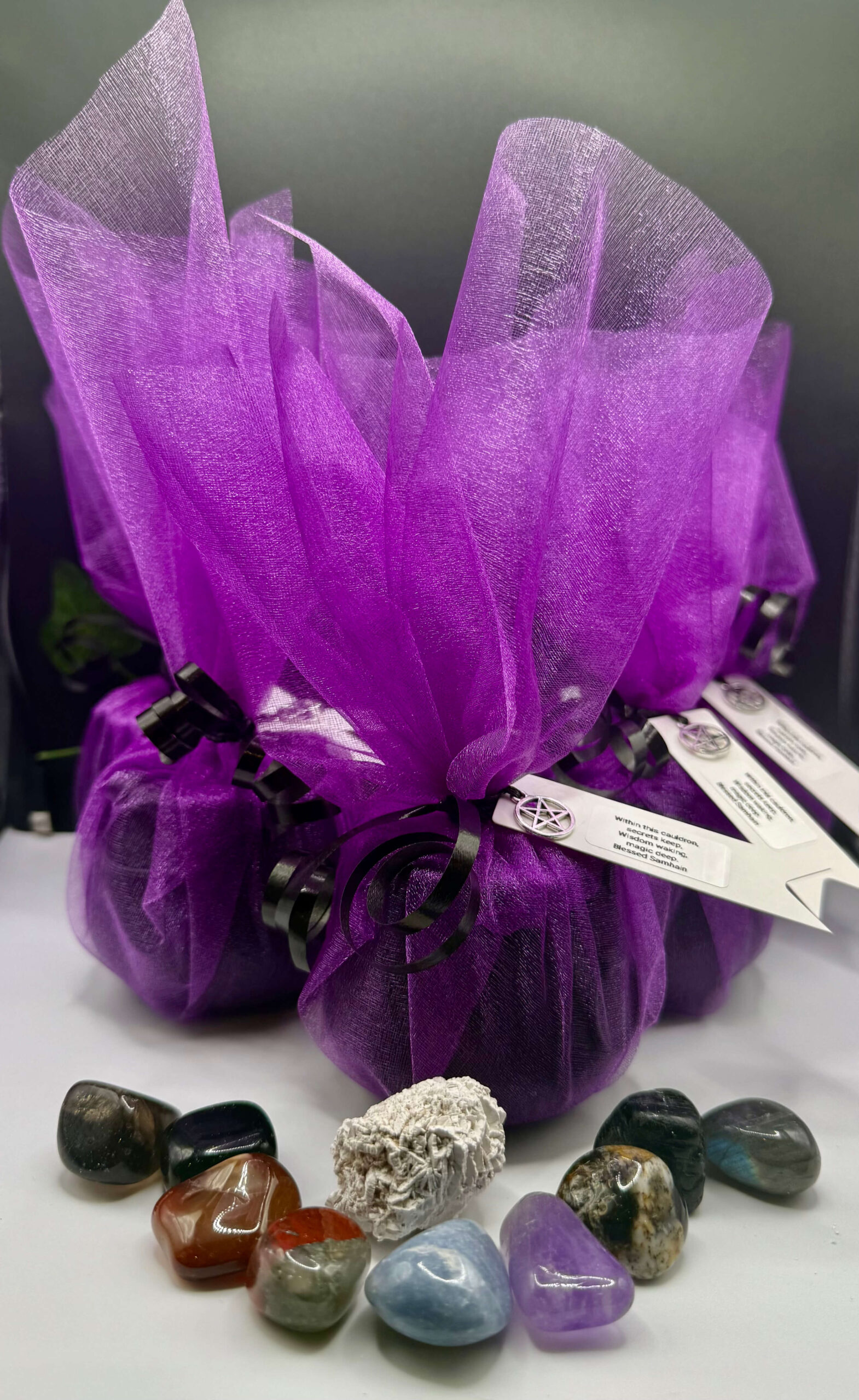 Violet gift wrapped in organza with black ribbon and pentagram charm, surrounded by polished healing crystals and stones, symbolizing energy work, spiritual healing, and crystal therapy at The Goddess Haven.