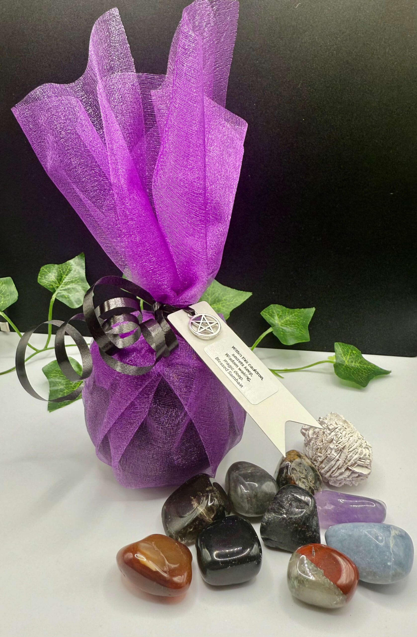Violet smudging bundle with crystals, including rose quartz, obsidian, and selenite, wrapped in purple organza and black ribbon, with a pentagram charm, on a white surface, ideal for spiritual cleansing and energy healing.