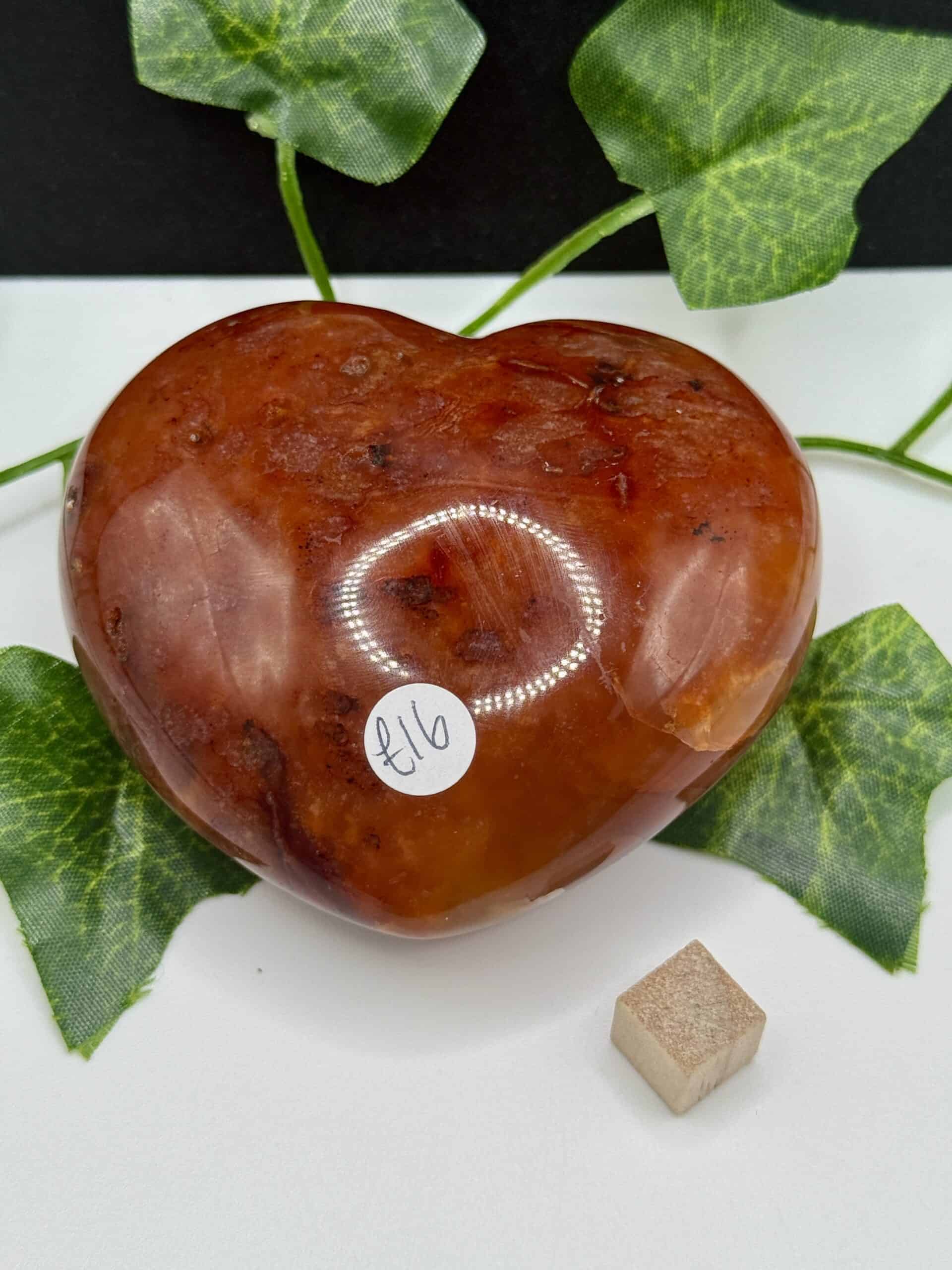 Polished heart-shaped orange calcite gemstone with price sticker, natural green leaves in the background, on a white surface, for healing, energy clarity, and spiritual wellness.