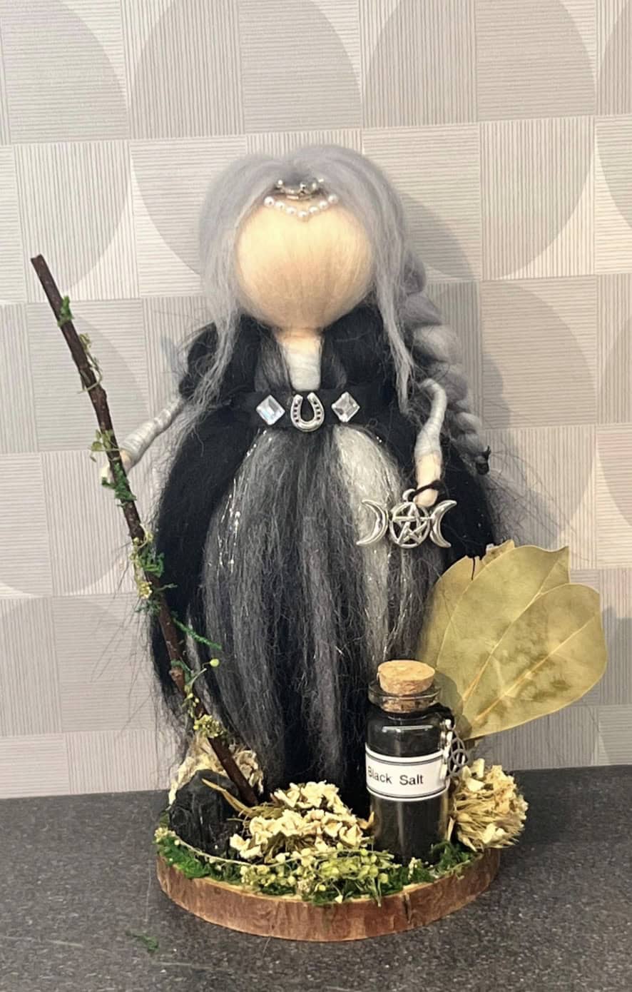 Enchanting witch doll with long gray hair, black dress, and mystical accessories, holding a twig and surrounded by herbs and natural elements. Perfect for spiritual or witchcraft-themed decor.
