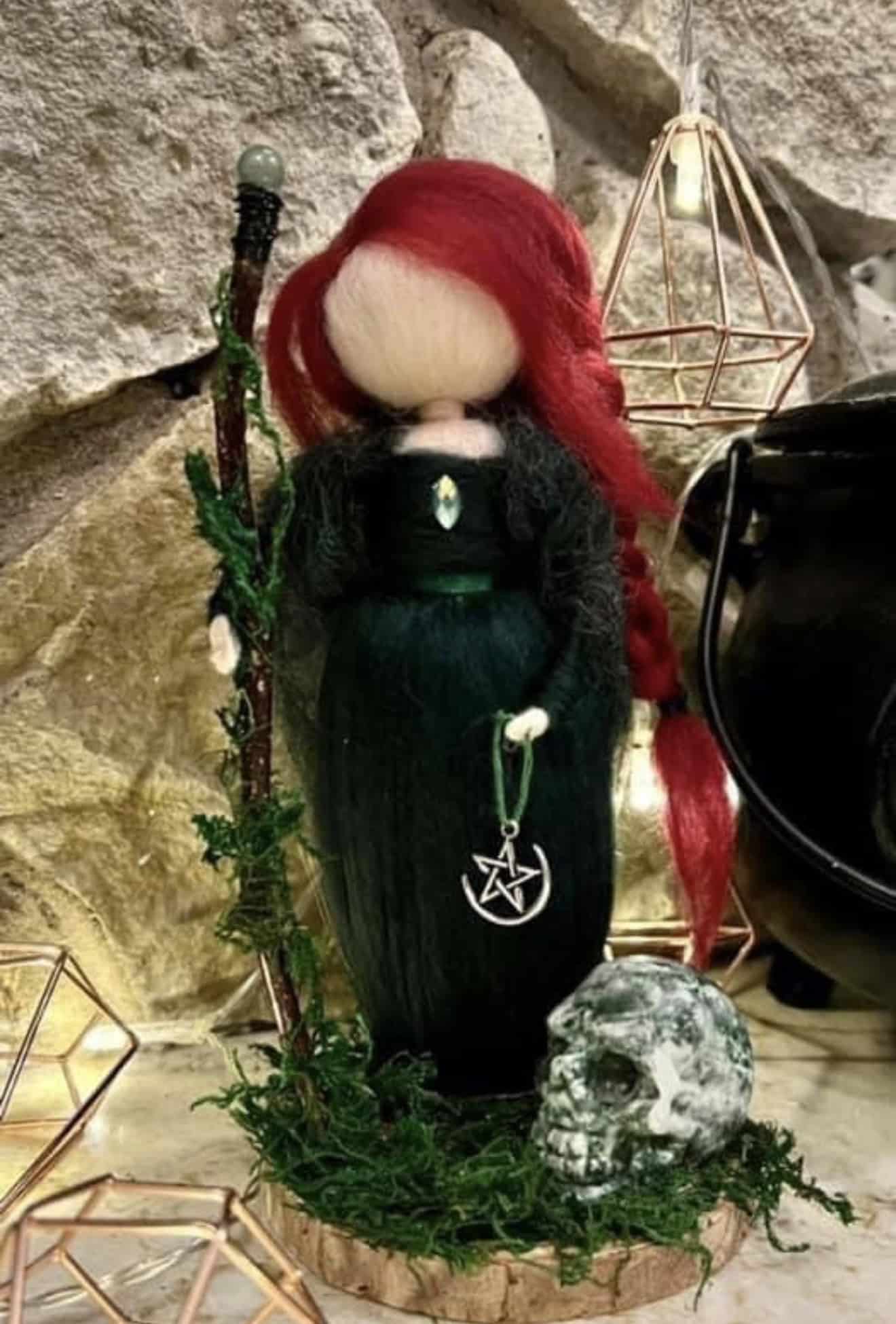 Handmade witch doll with long red hair, black dress, and mystical accessories, displayed with skull and greenery for Halloween or spiritual decor.