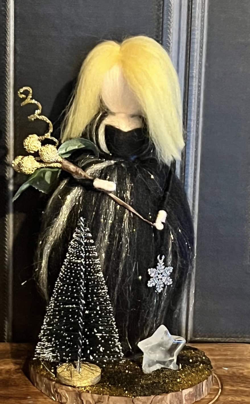Enchanted witch doll with blonde and black hair, dressed in a black cloak, holding a small broomstick, and decorated with a snowflake pendant. Perfect for Halloween or winter-themed decor.