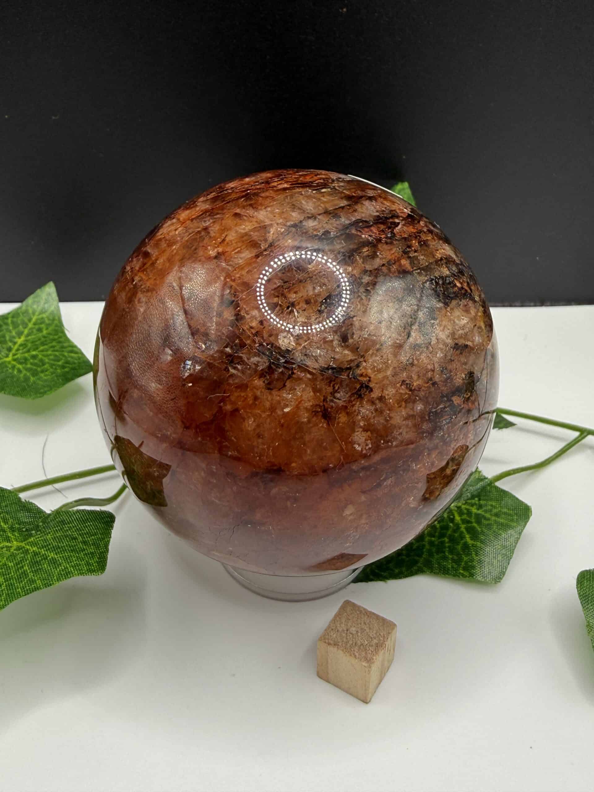 Brown gemstone crystal sphere with polished surface, surrounded by green ivy leaves and a small wooden block, on a white and black background.