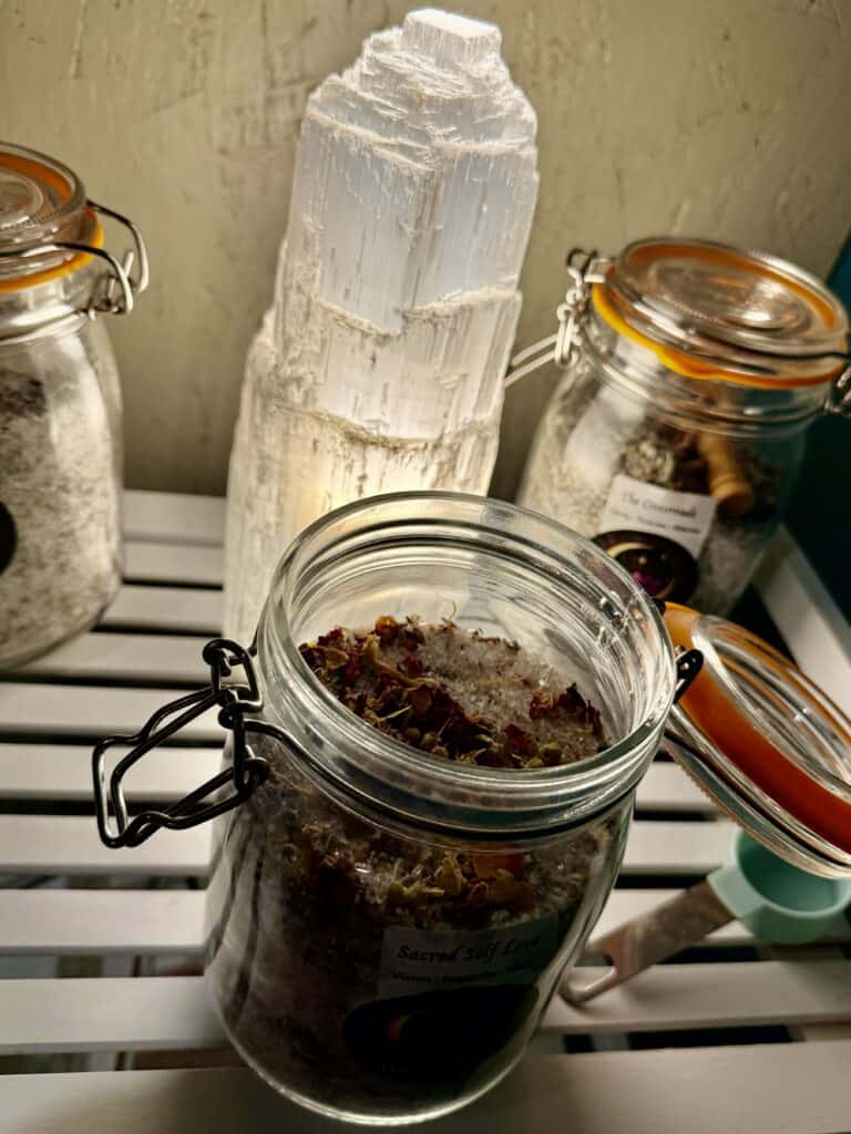Close-up of a healing crystal and herbal jars on a wooden surface, featuring a large white selenite crystal and various herbal infusions in glass jars, ideal for spiritual wellness.
