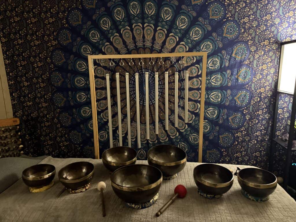 Calming sound healing room featuring seven crystal singing bowls arranged on a soft mat, with a decorative mandala wall art in the background, creating a tranquil atmosphere for relaxation and meditat.