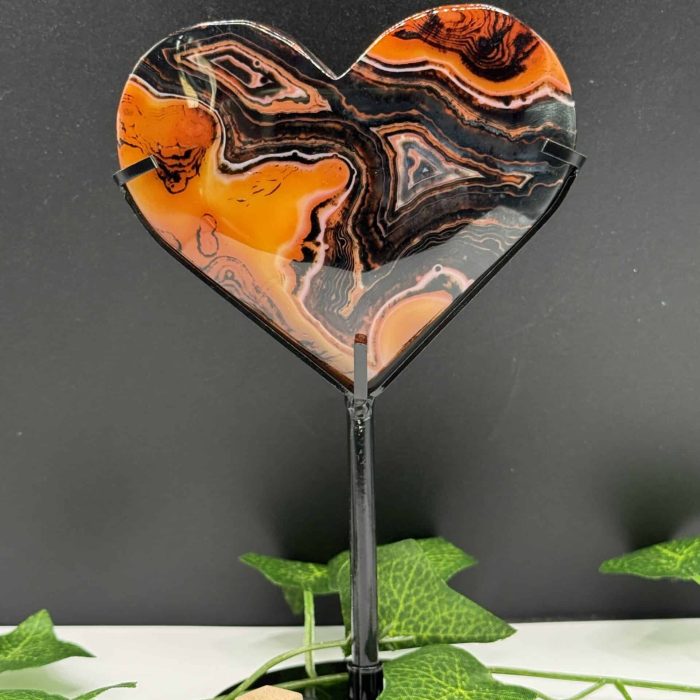 Heart-shaped agate crystal ornament with vibrant orange, black, and white banding, mounted on a sleek black stand, surrounded by green ivy leaves on a white surface against a dark background.